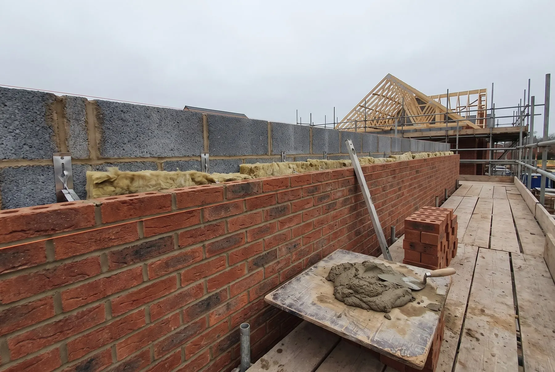 A cavity wall going up: facing brick, blockwork, wall ties and insulation, with a mortar board on the scaffold