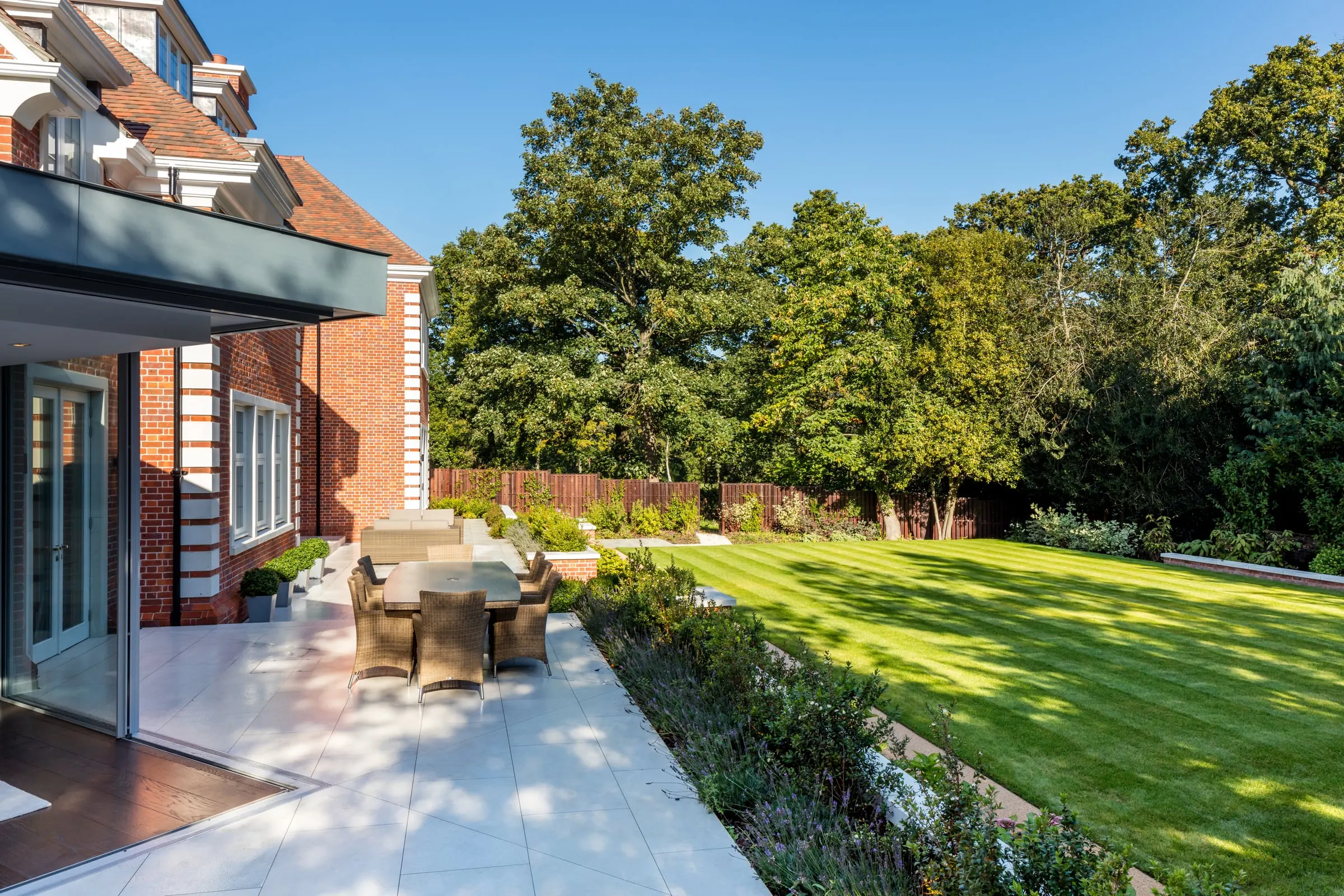 Stone terrace with dining furniture beside a brick house, with a planted border, lawn and mature trees
