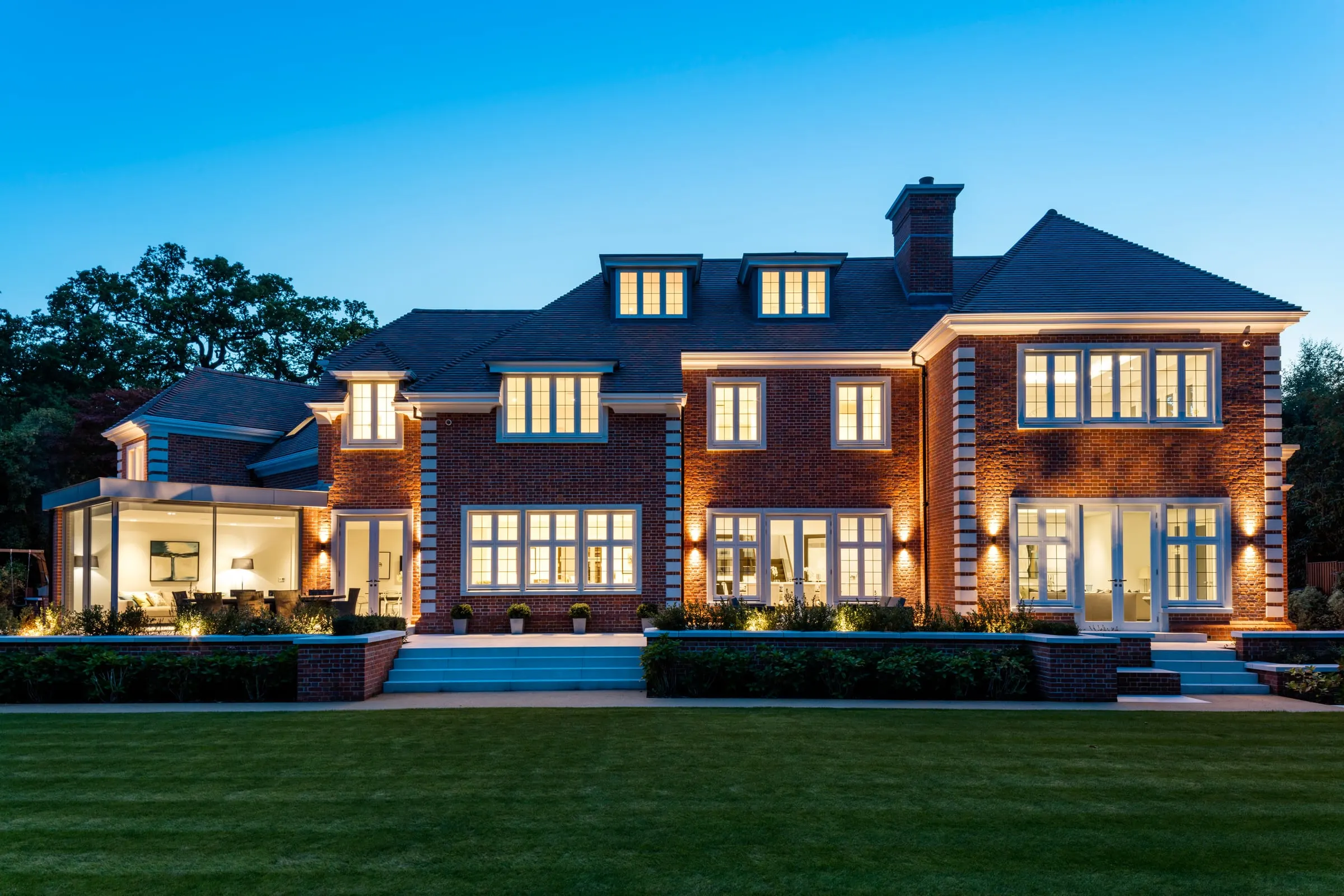 The back of a large brick house at dusk, seen across its lawn with every window lit