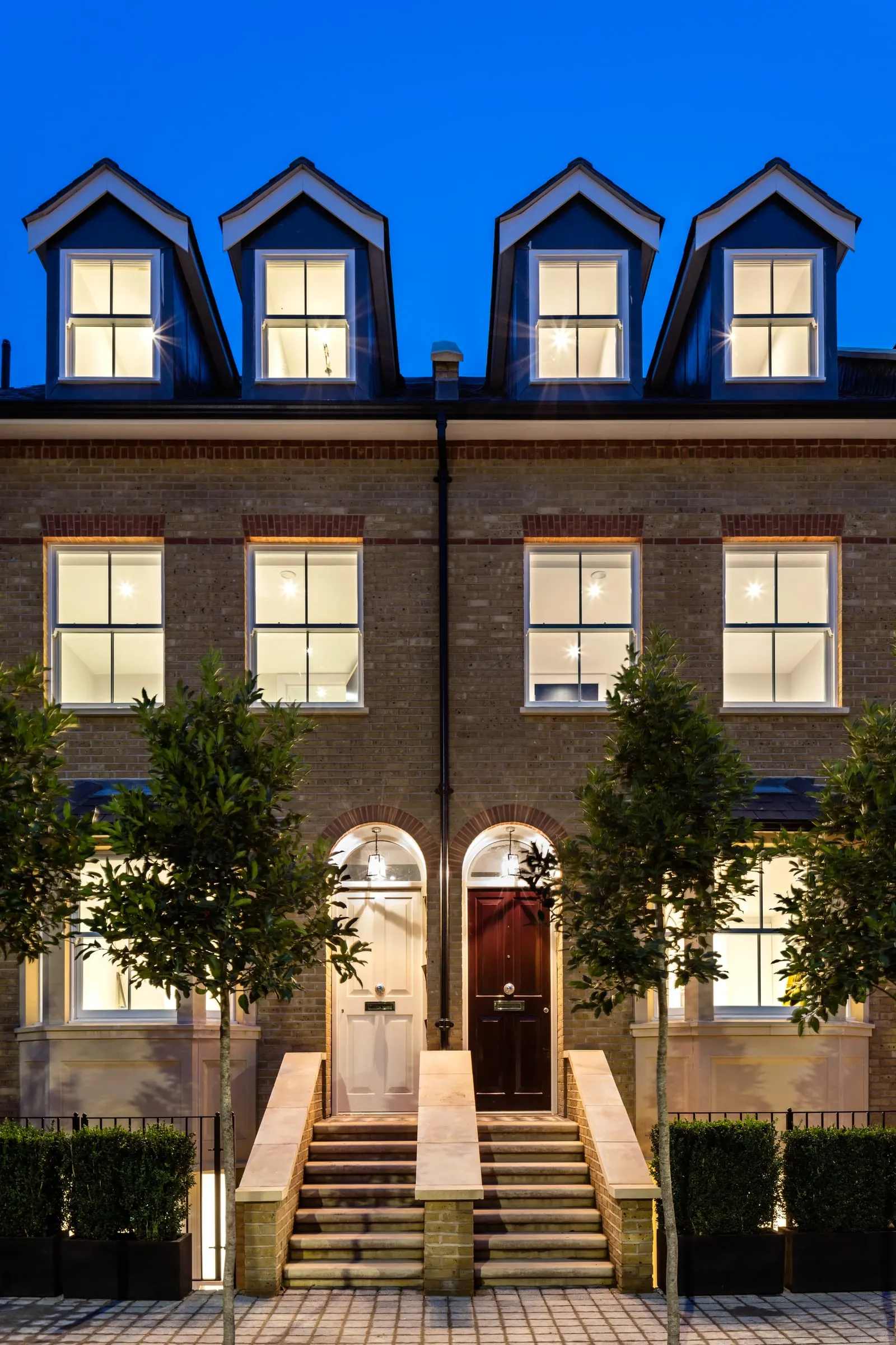 Two front doors, steps and bay windows of finished town houses, lit at dusk, Teddington